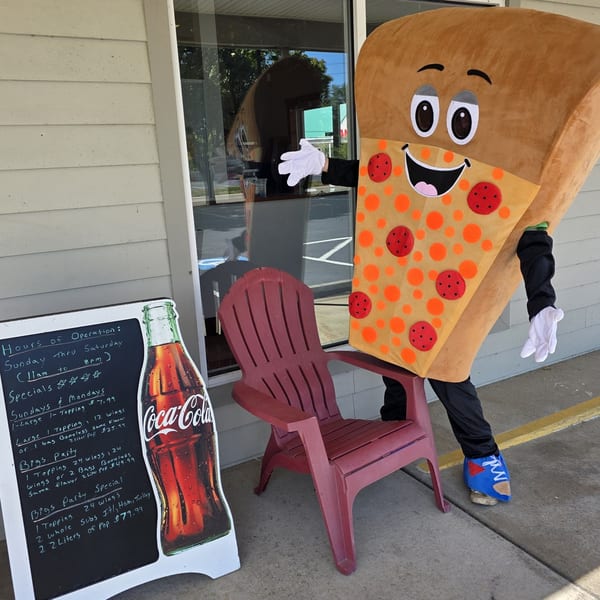 Big's Pizza mascot, a smiling pizza slice, stands next to a chalkboard sign with specials.