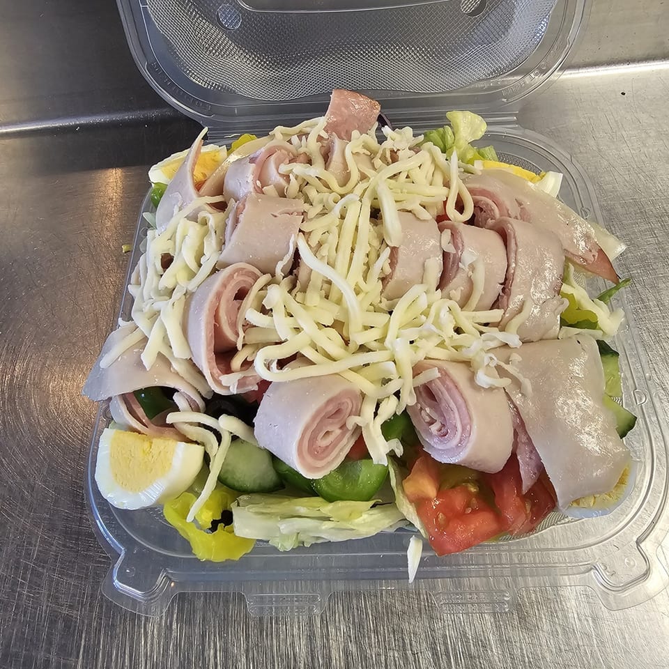Chef salad with rolled ham, turkey, shredded cheese, hard-boiled egg, and vegetables in a plastic container.