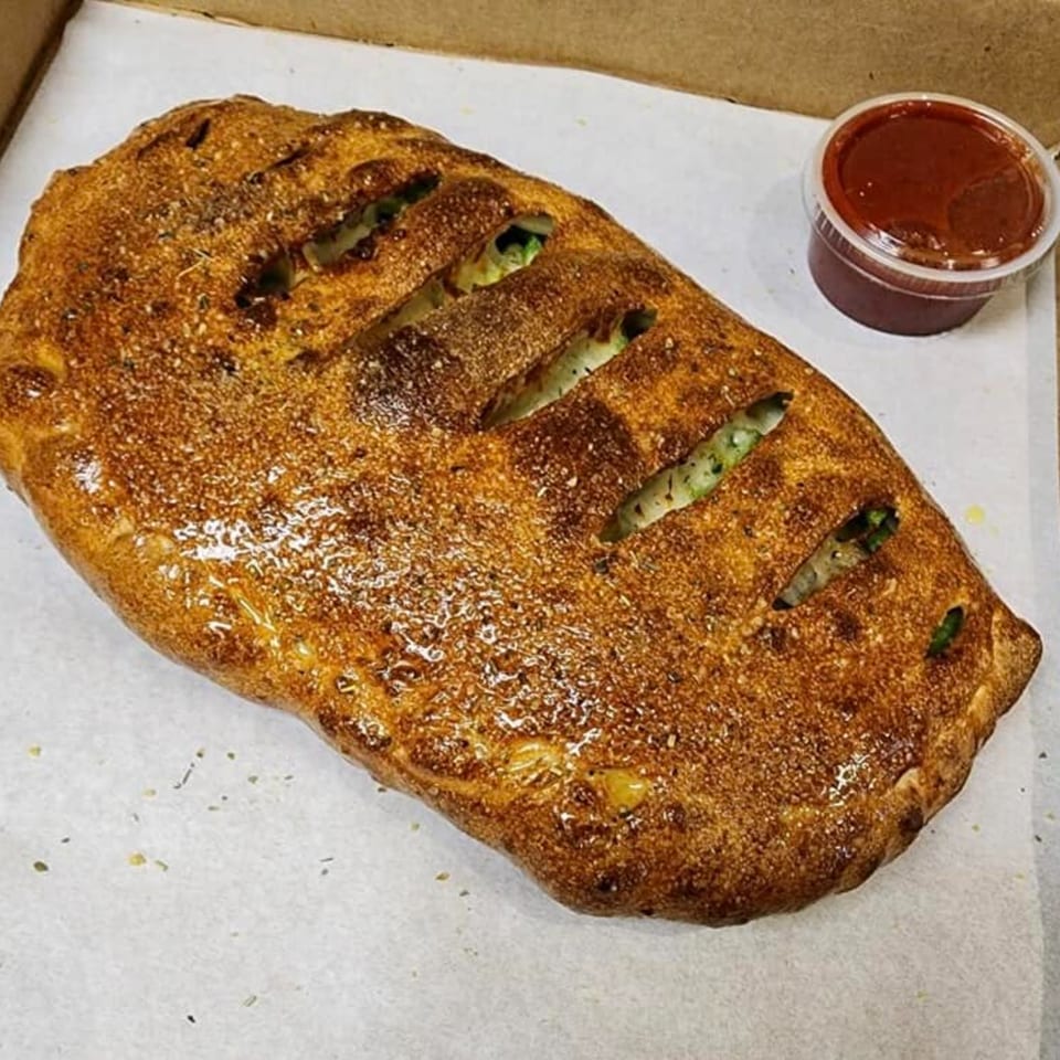 A golden-brown calzone with visible green filling and a side of red sauce in a take-out box.