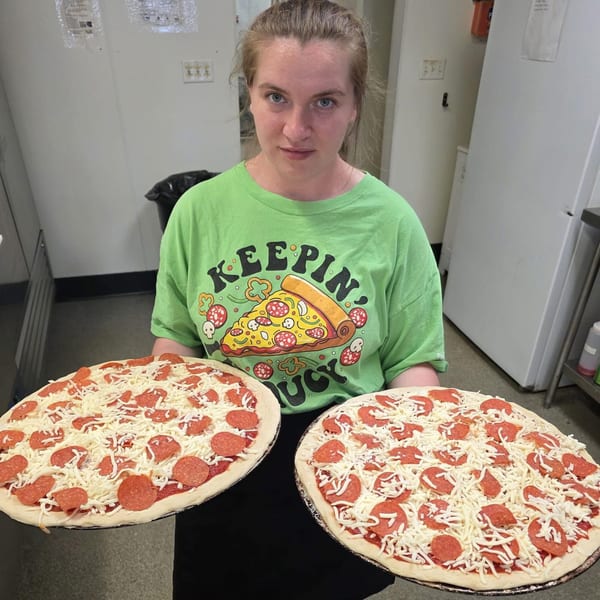 A Big's Pizza employee holds two uncooked pepperoni and cheese pizzas in a kitchen.