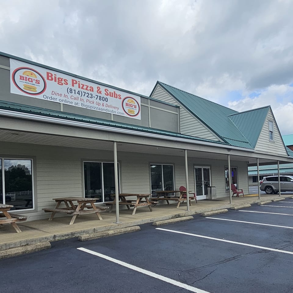 Exterior of Big's Pizza & Subs with a banner sign, picnic tables, and a parking lot under a cloudy sky.