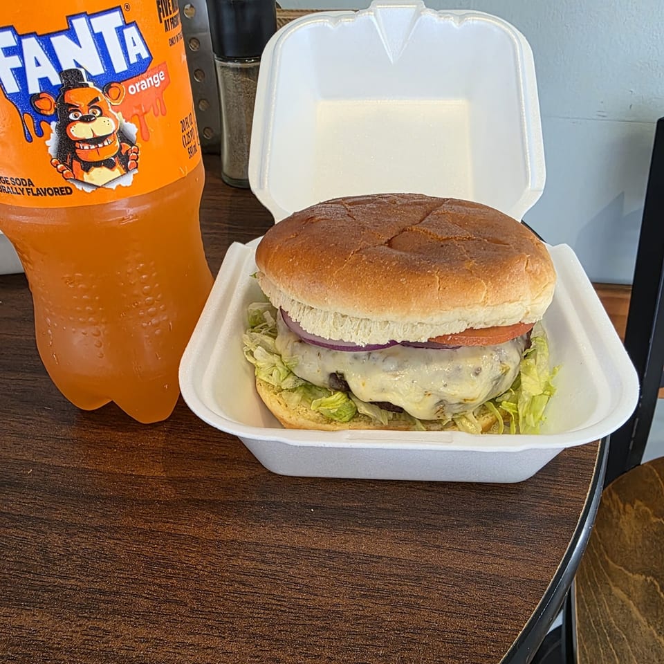 A cheeseburger with lettuce, tomato, and onion in a takeout container next to an orange Fanta bottle.